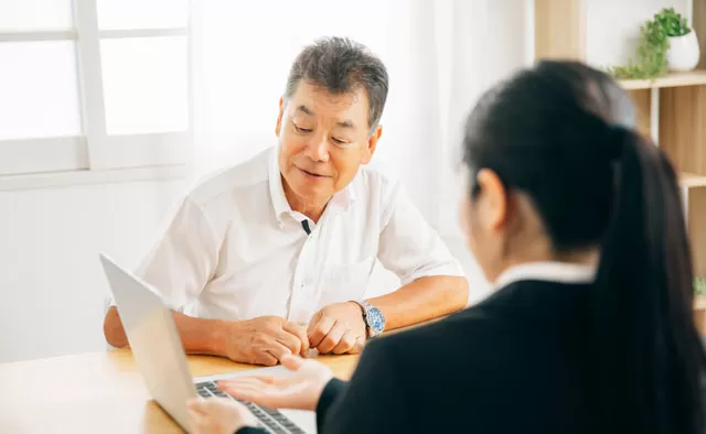 白いシャツを着た中年男性がテーブル越しにスーツ姿の女性と対面して話している場面